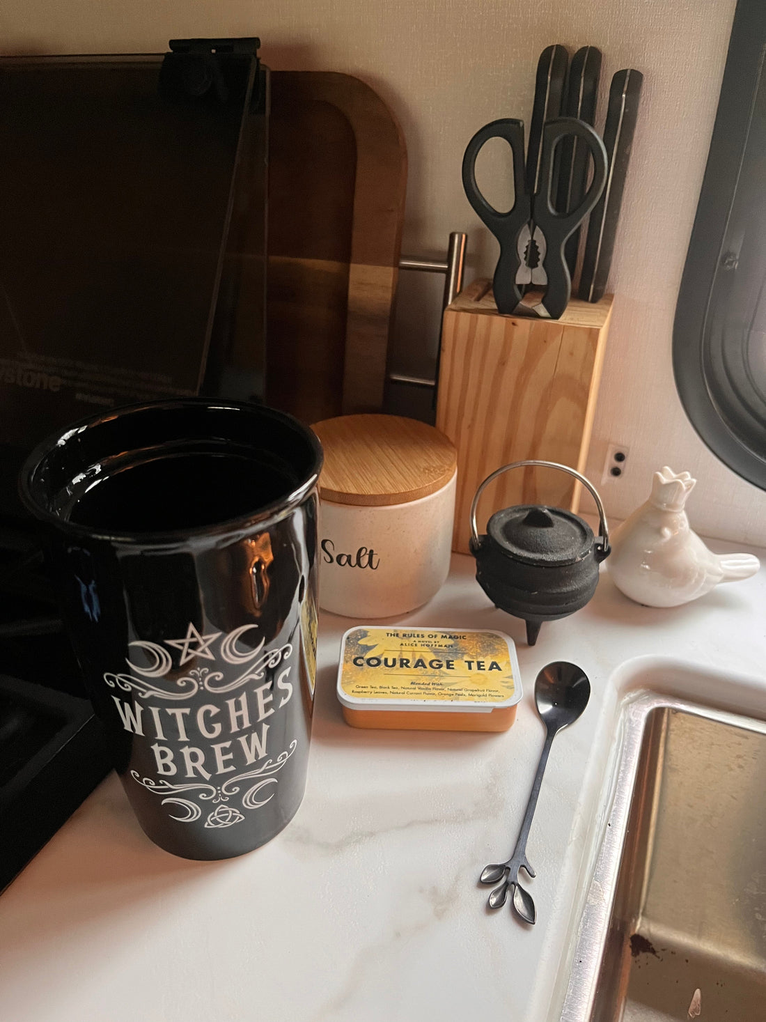 A cozy kitchen counter with a Witches Brew mug, Courage Tea tin, mini cauldron, and salt jar on a rainy morning
